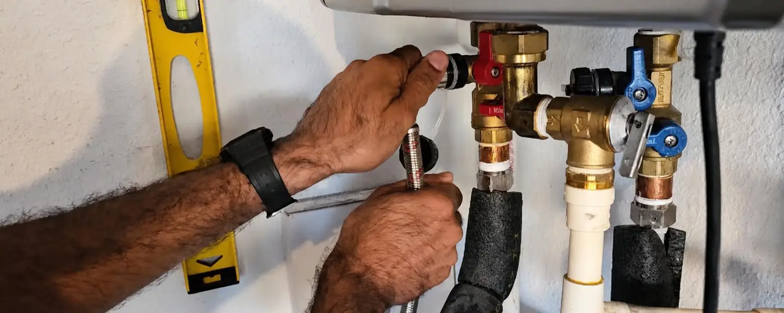 Hands adjusting valves under a wall-mounted tankless water heater with a level resting on the pipes